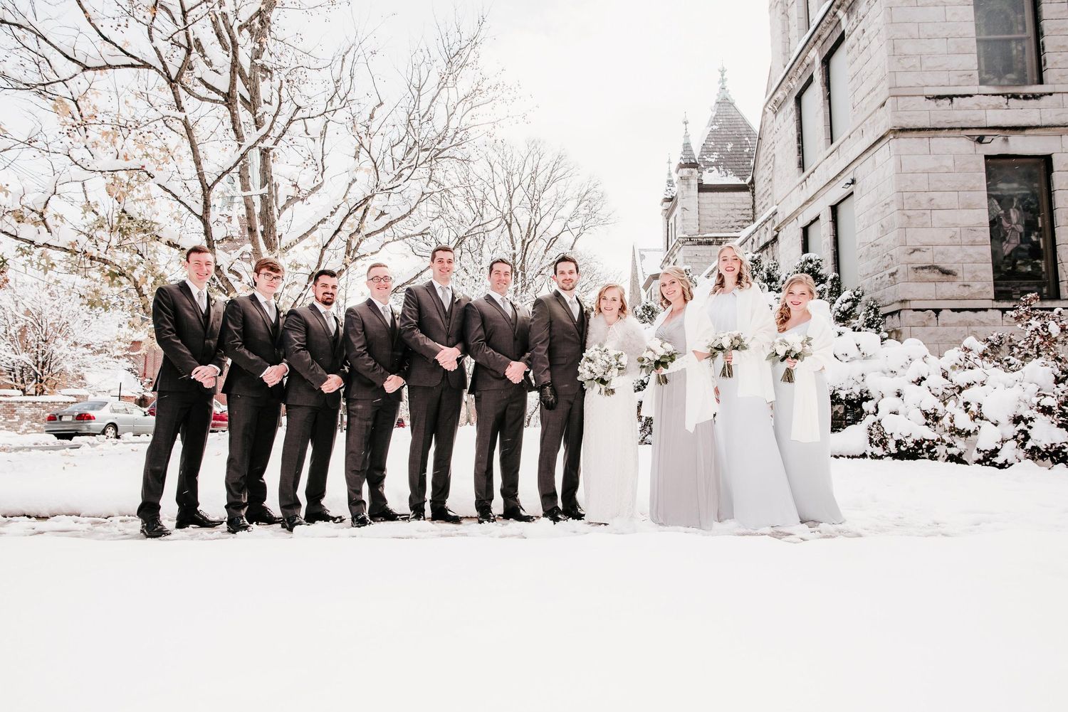 Wedding party poses in snow outside historic building, groomsmen in dark suits beside bridesmaids in grey gowns with bride in white.