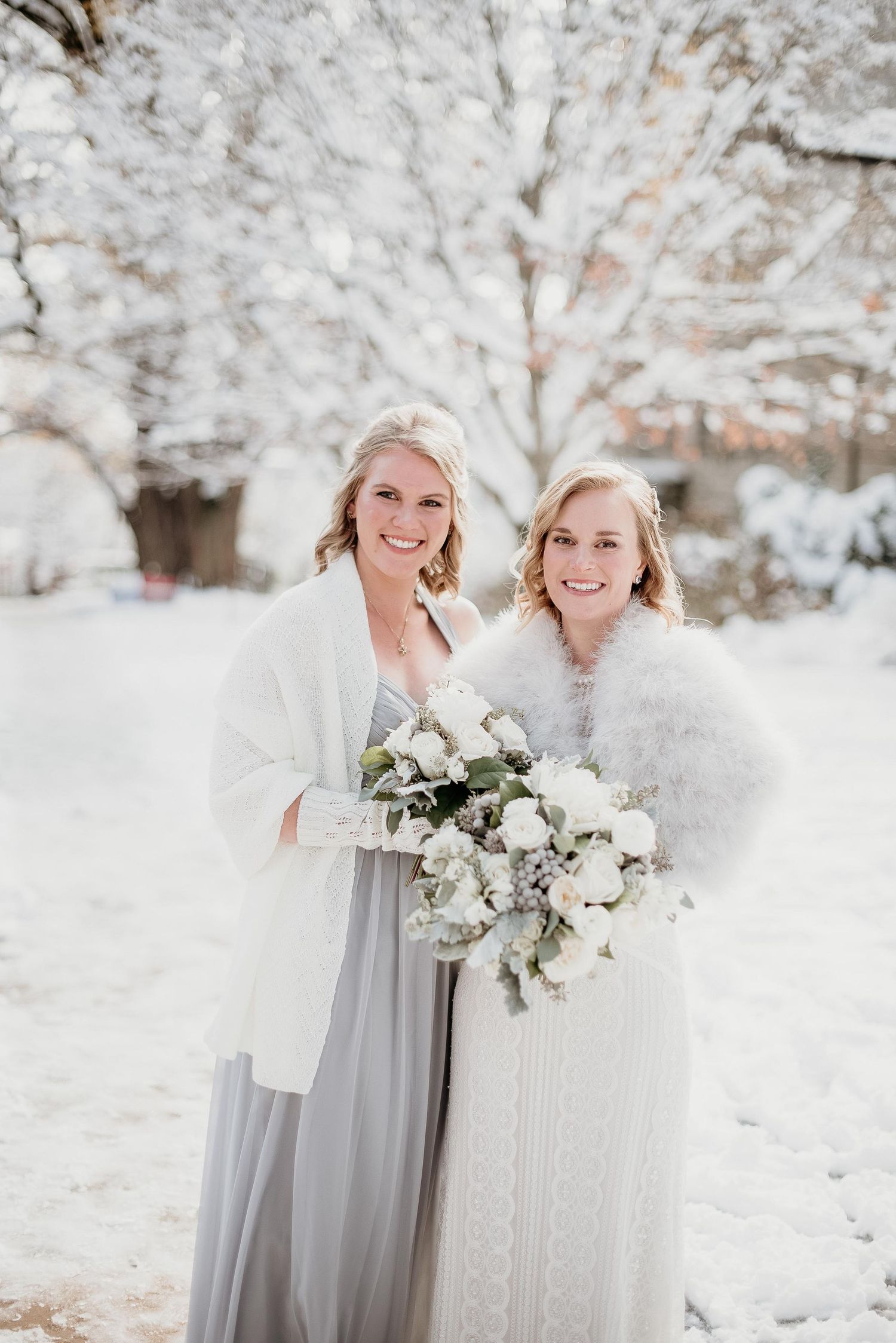 Two brides in white gowns and fur wraps hold white floral bouquets outdoors in a snowy winter wedding setting.