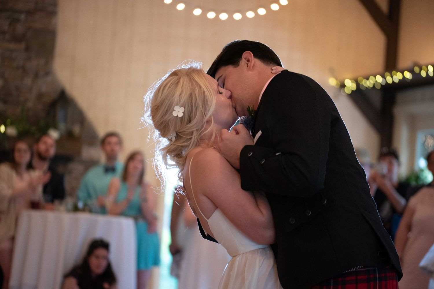 Bride and groom share first dance kiss at wedding reception in rustic venue with string lights.