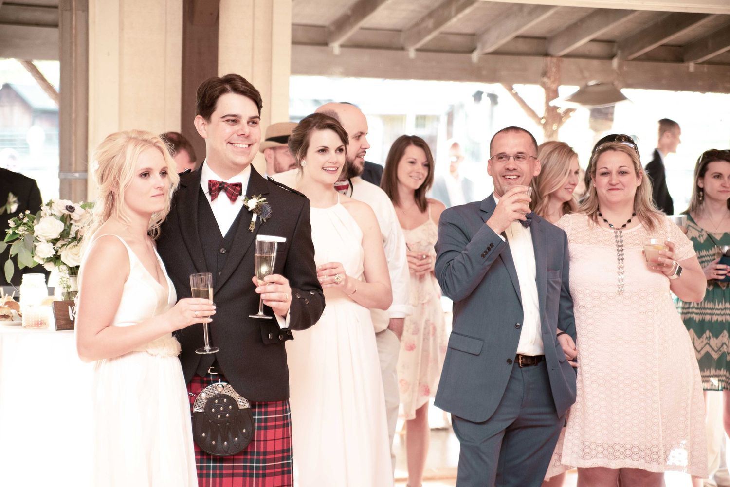 Wedding party toasting with champagne, groom in tartan kilt beside bride in white dress, surrounded by guests.