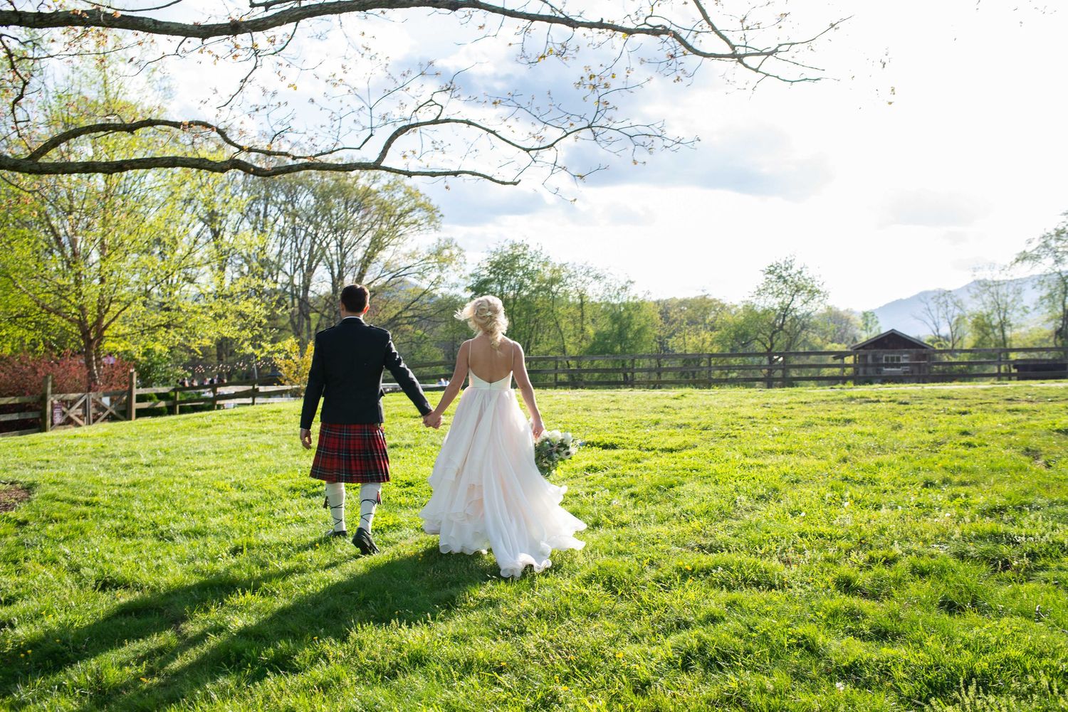 Bride and groom walk hand-in-hand across lush green field, groom wearing traditional Scottish kilt at outdoor wedding.