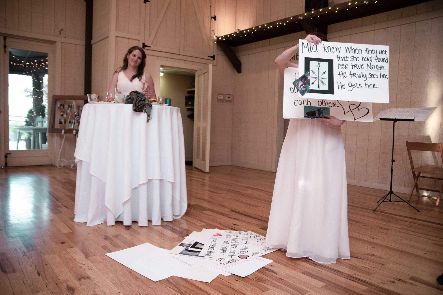 Bride stands at white cocktail table while bridesmaid holds handwritten sign during indoor wedding reception on hardwood floors.