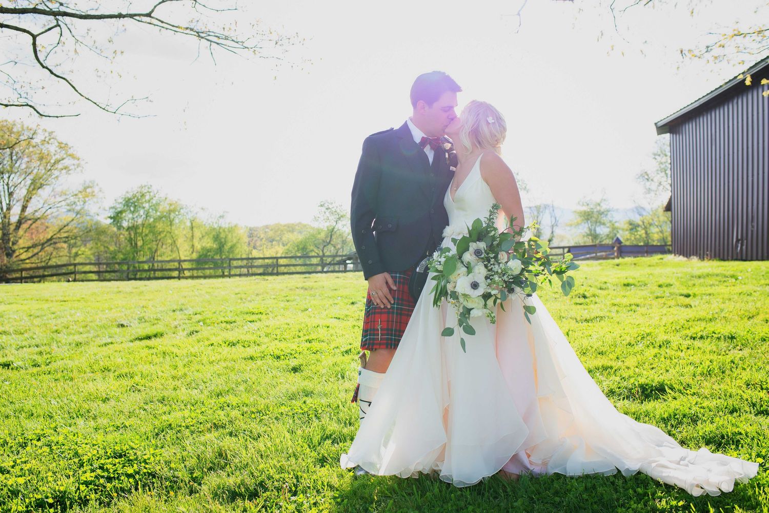 Bride and groom share a romantic moment in a sunlit green field, with bride holding a lush bouquet near a rustic barn.