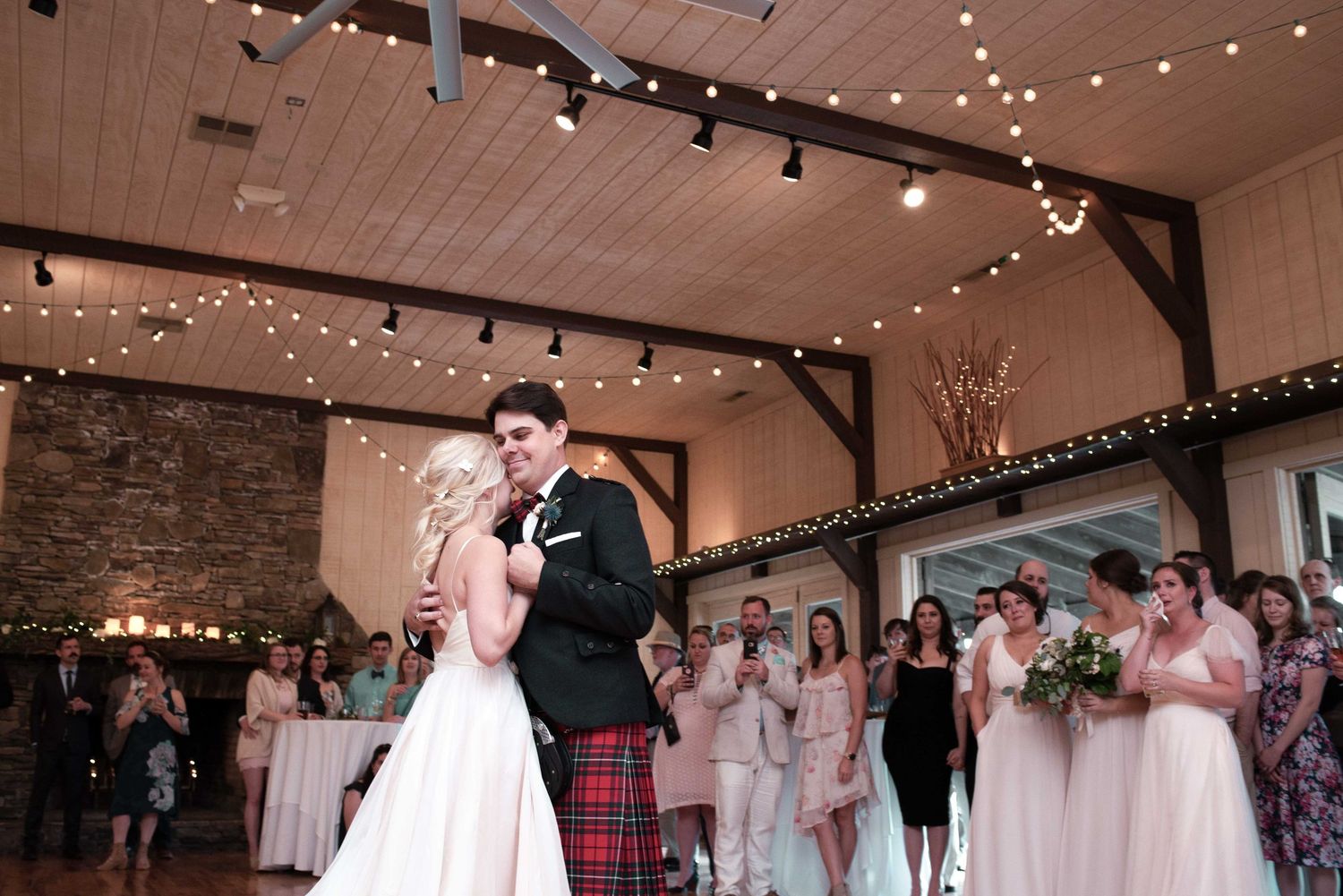 Bride and groom share first dance in rustic barn venue with fairy lights, surrounded by bridesmaids in pink dresses.