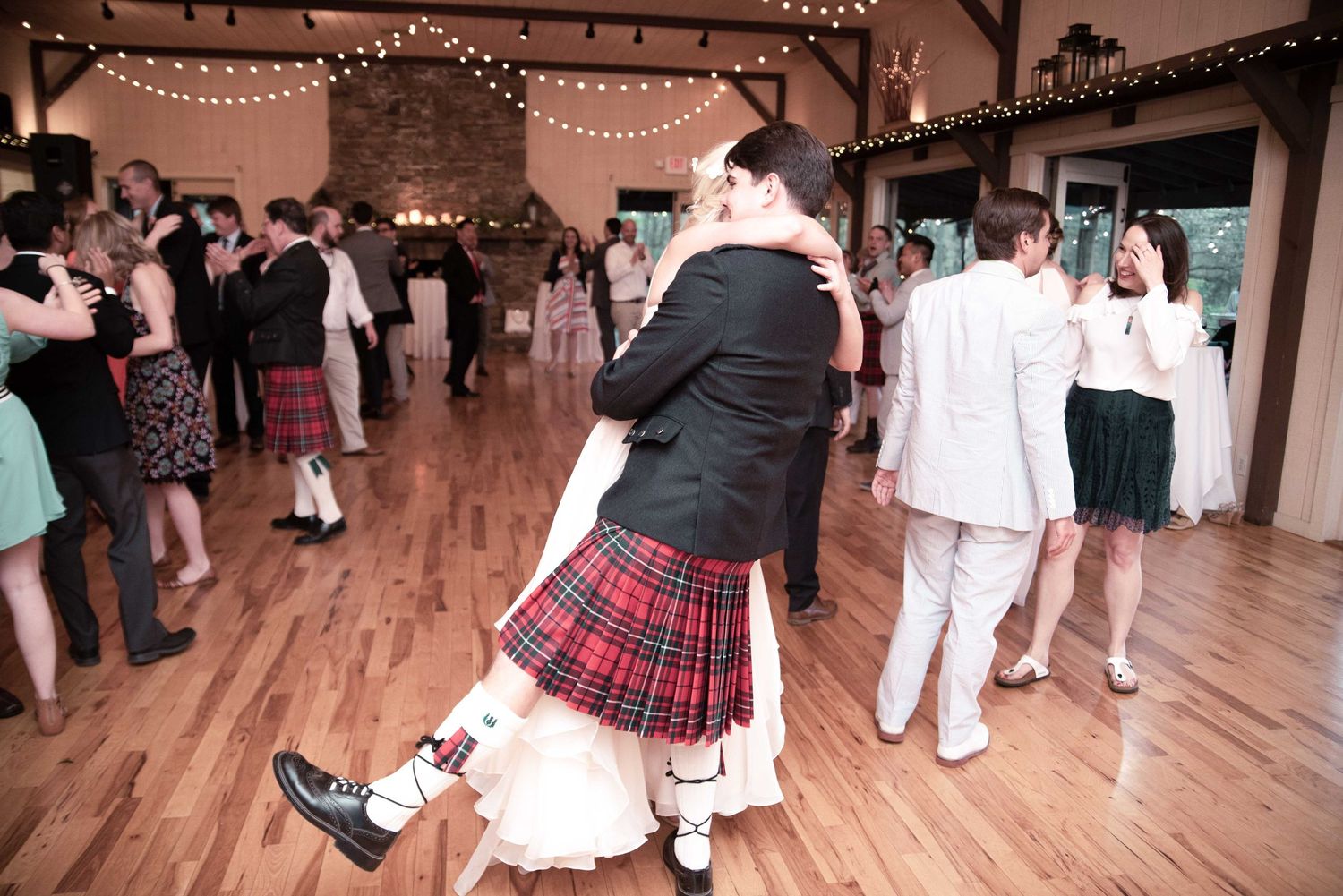 Couple sharing romantic dance at wedding reception, groom wearing traditional Scottish kilt on wooden dance floor.