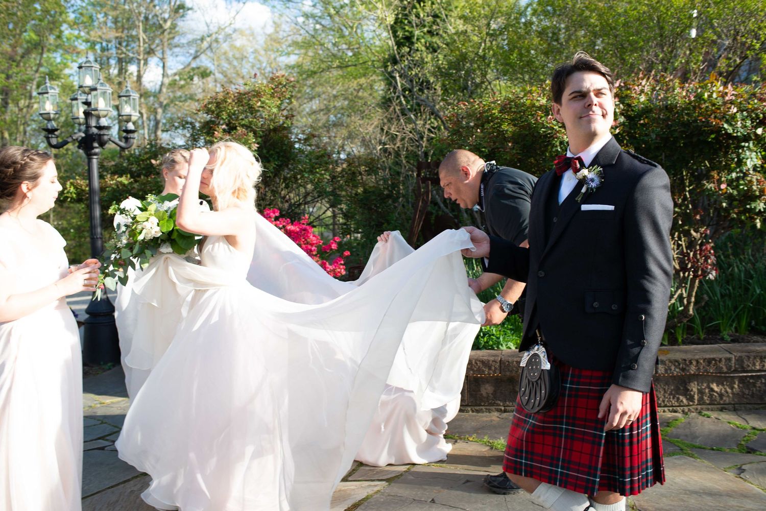 Groom in red tartan kilt and black jacket smiles as bride in white gown holds bouquet at outdoor wedding ceremony.