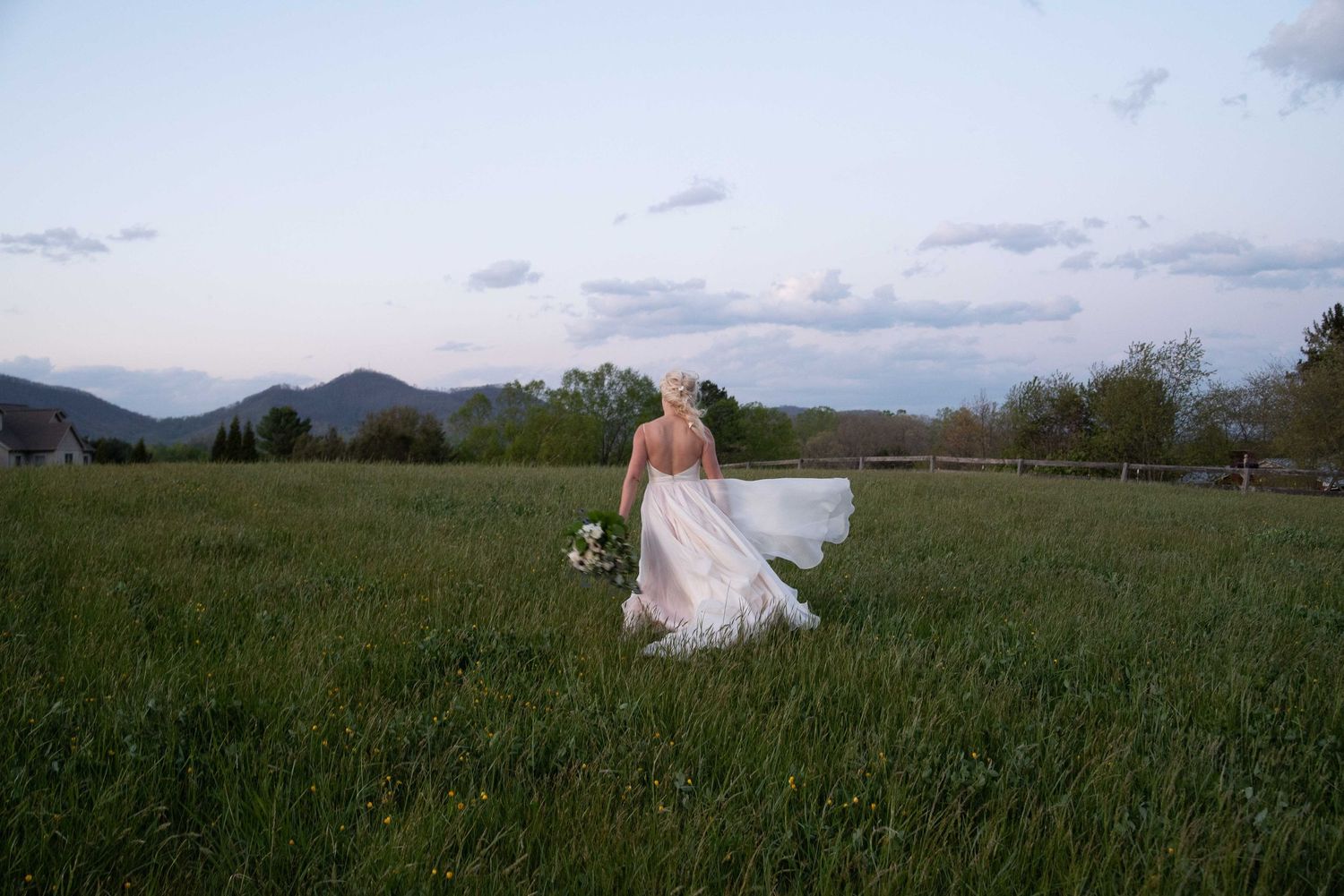 Elegant bride walking through lush green meadow in backless wedding gown with mountains at sunset.