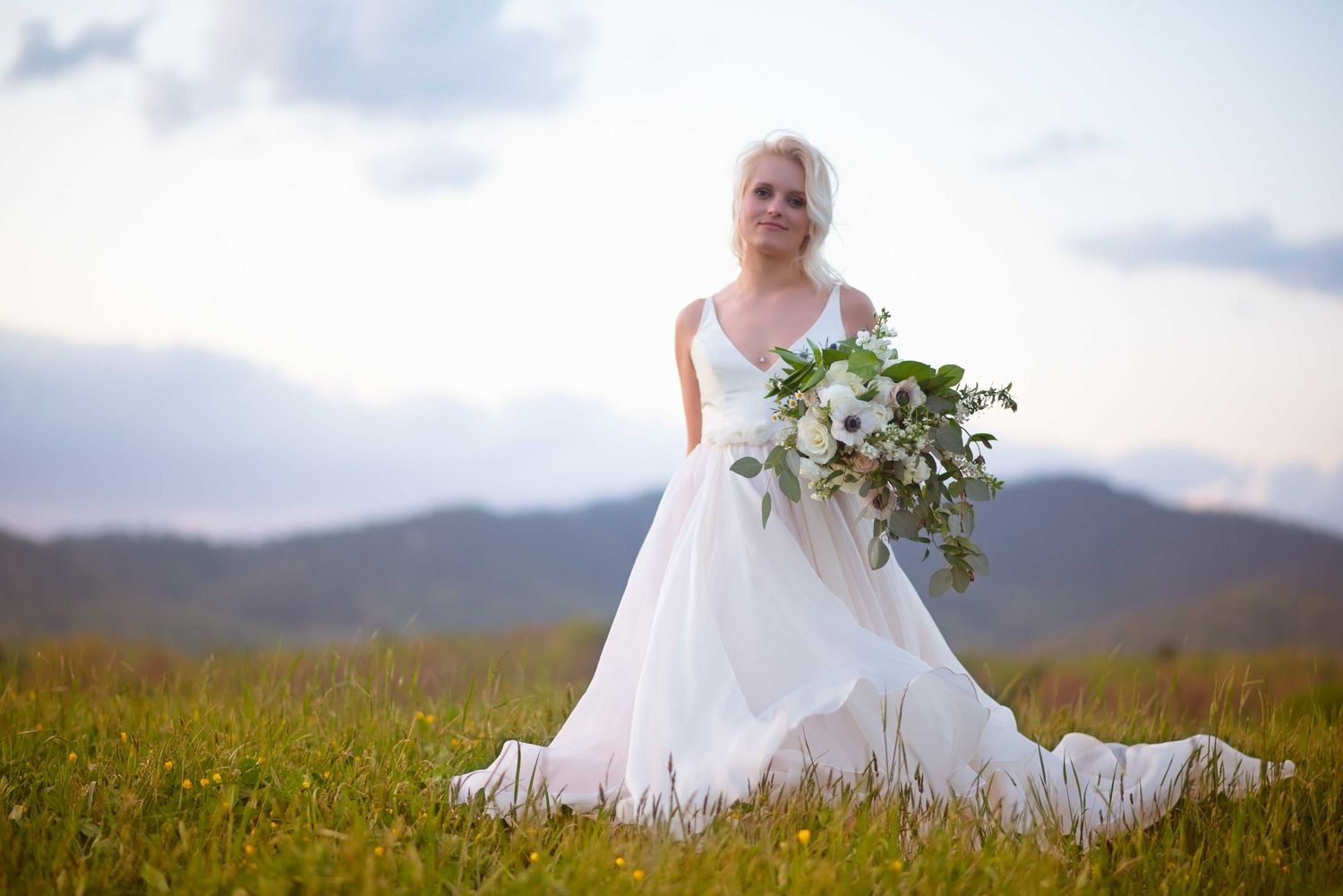 Bride in white gown holding green and white bouquet stands in mountain meadow under cloudy sky.