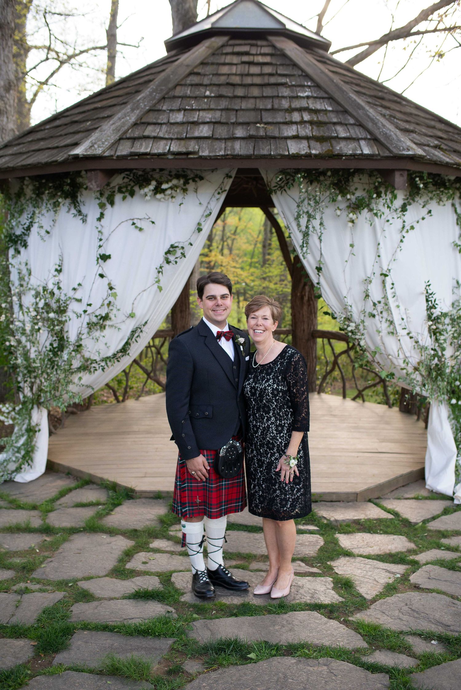 Couple posing in front of a decorated gazebo with white drapes and greenery, man wearing a red tartan kilt at an outdoor wedding.