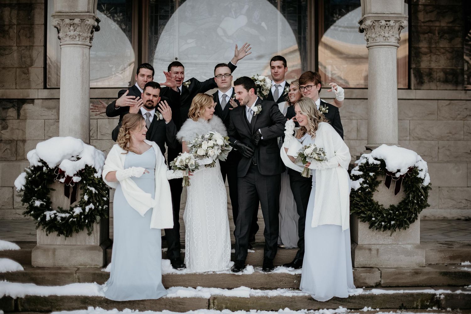 Wedding party poses on snowy church steps in winter, bridesmaids in white gowns, groomsmen celebrating behind bride and groom.