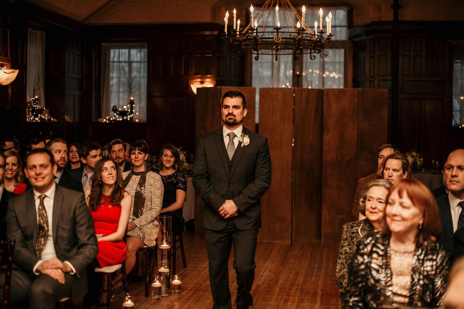 Groom standing at wedding altar in dark suit, guests seated in elegant wood-paneled venue with chandelier lighting.