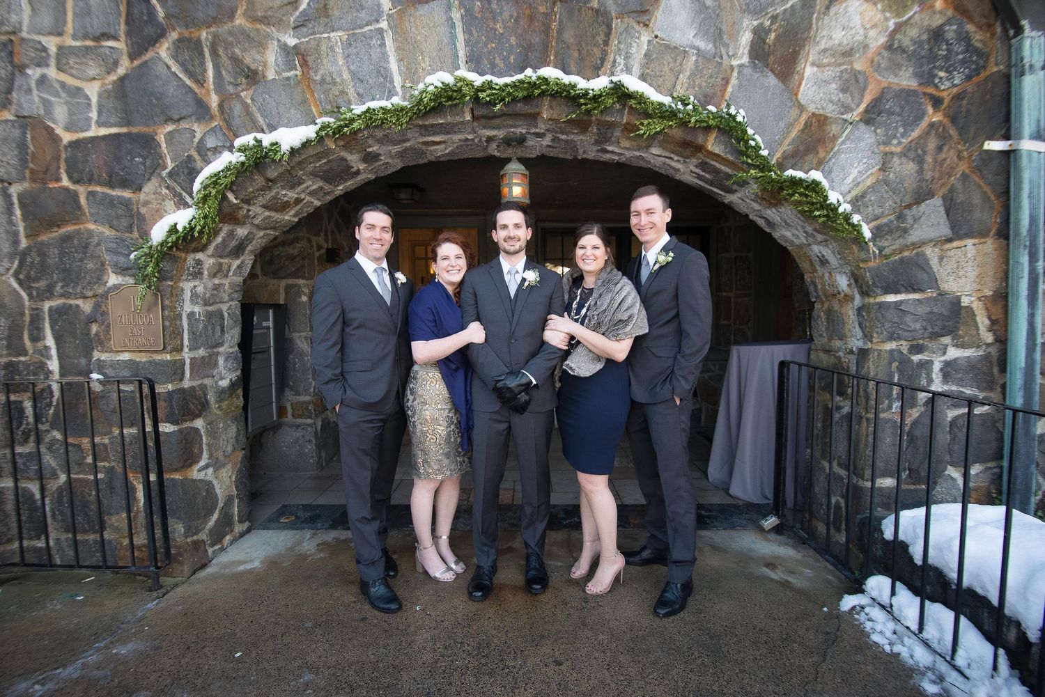 Five elegantly dressed guests posing under a stone archway decorated with greenery at a winter wedding venue.