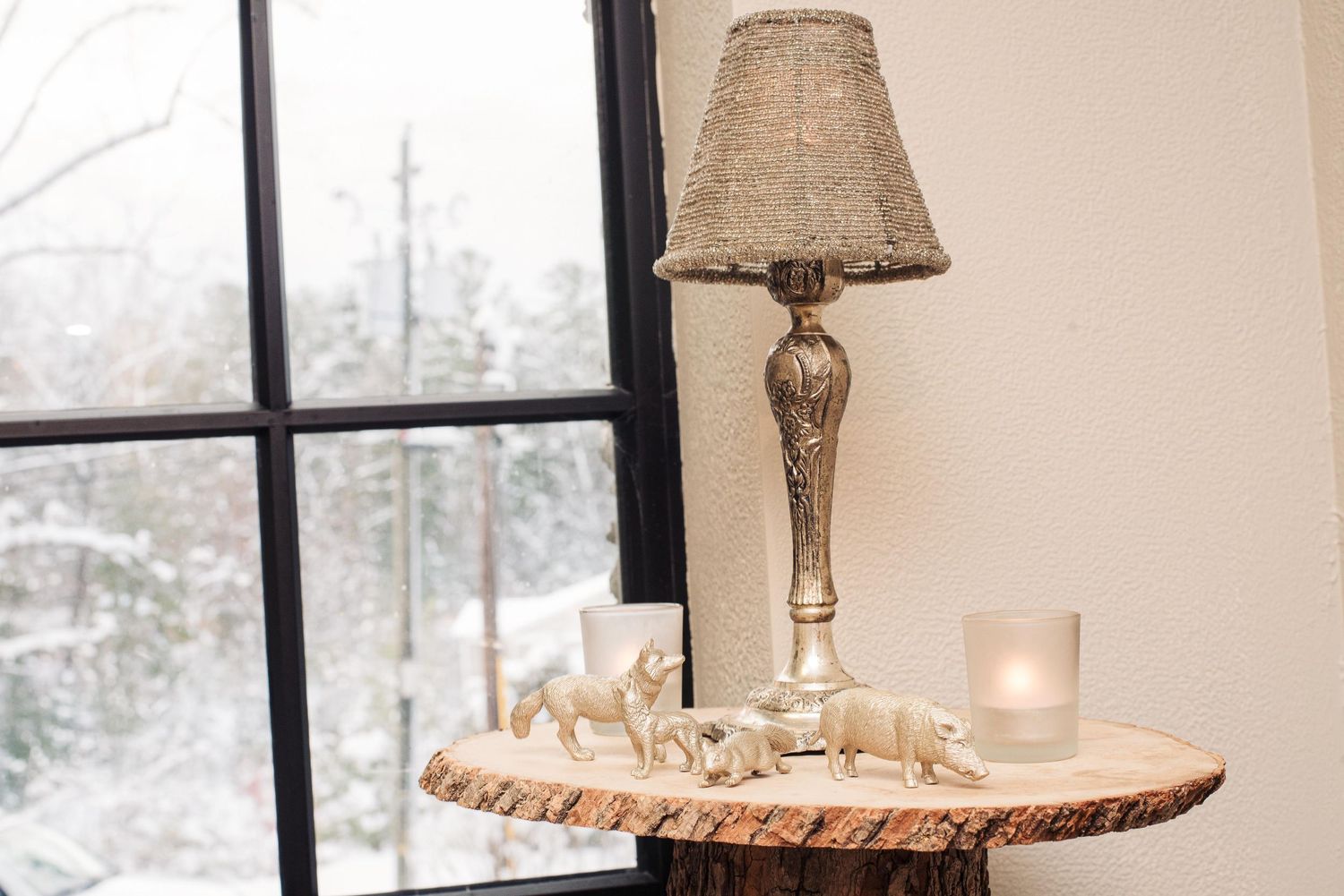Rustic wood slice table with silver lamp, white candle, and animal figurines beside a snowy winter window.
