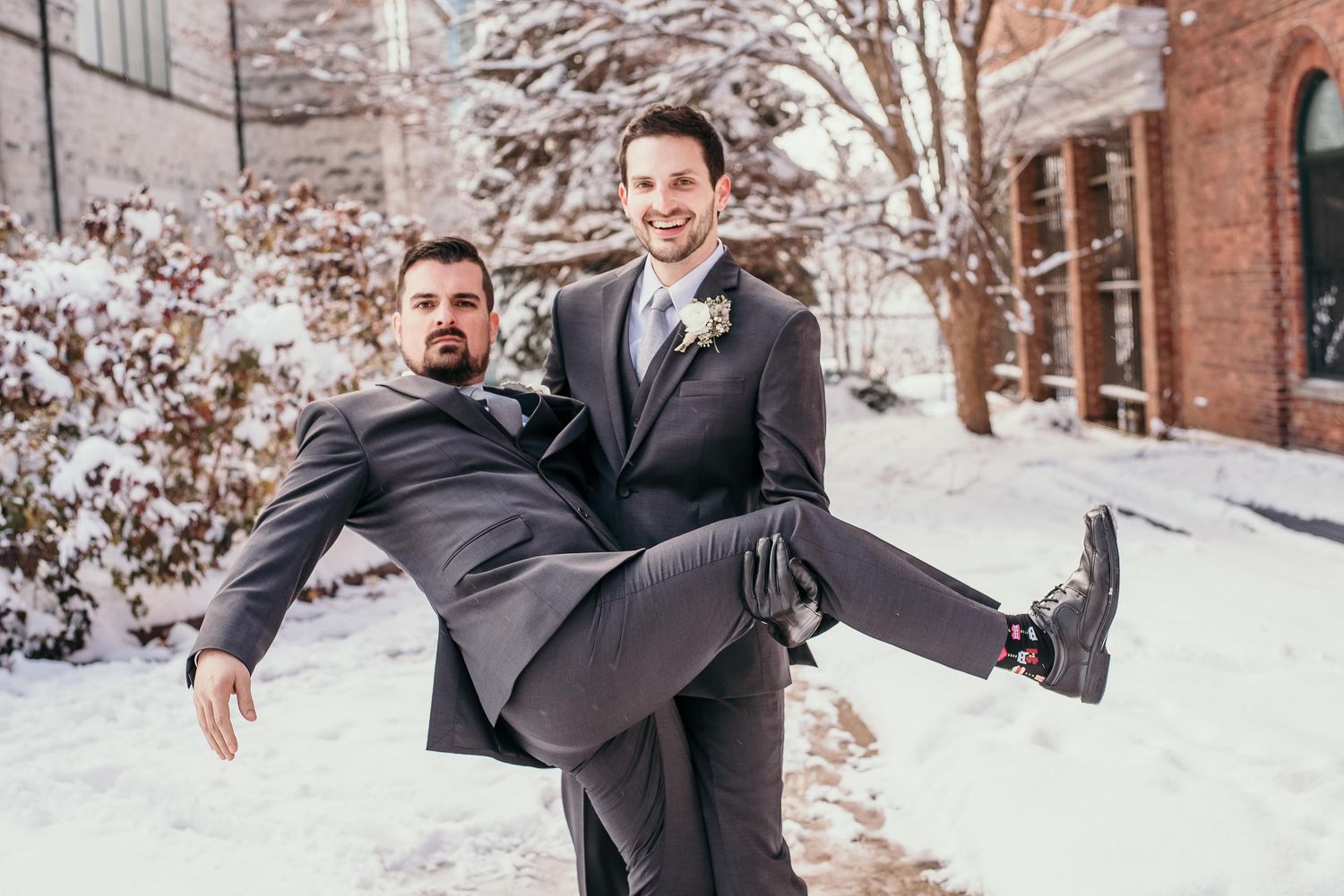 Two grooms in gray suits pose playfully in snowy winter setting, one lifting the other during wedding photo.