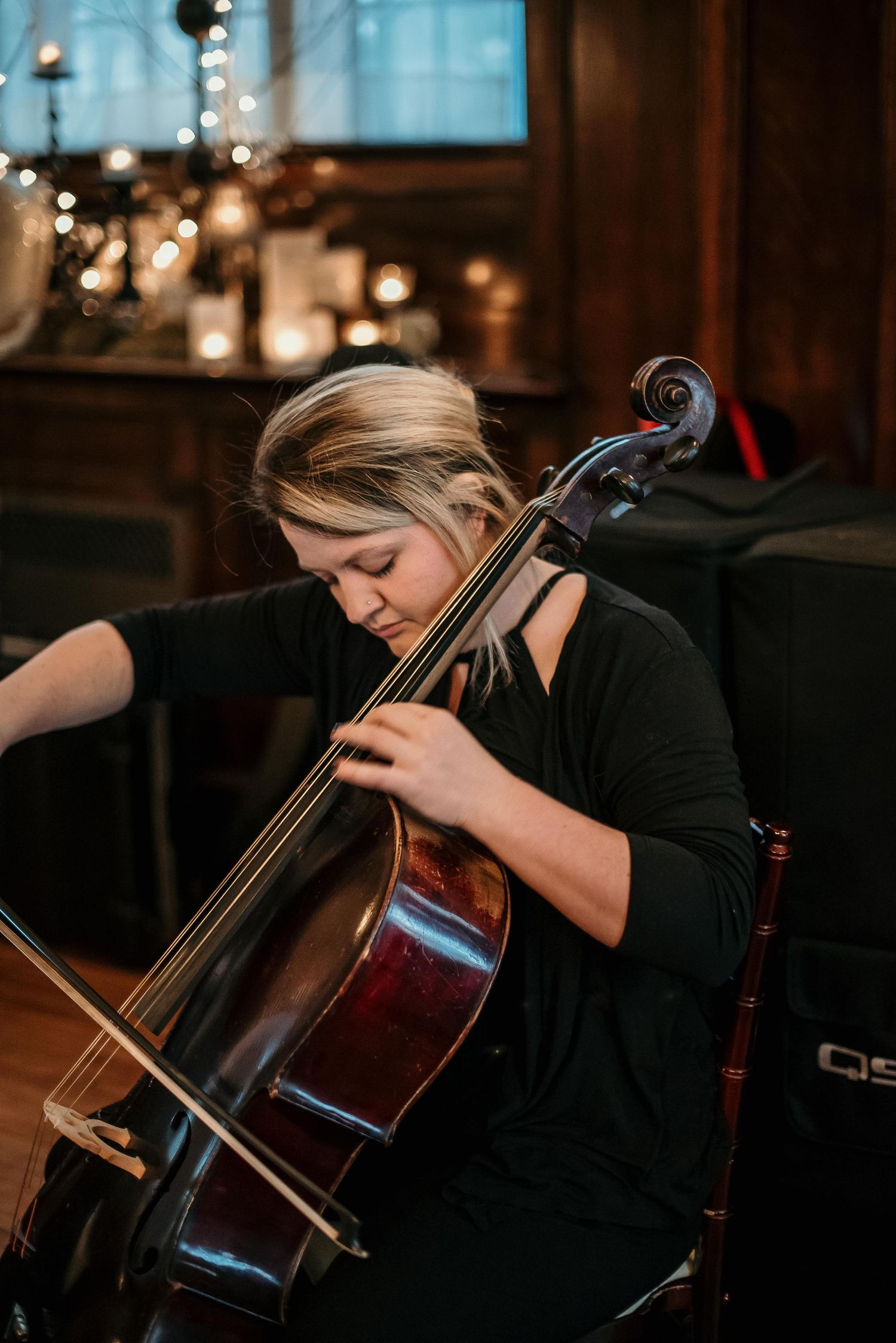 A blonde woman in black dress plays cello in a warmly lit room with candles and bokeh lights in background.