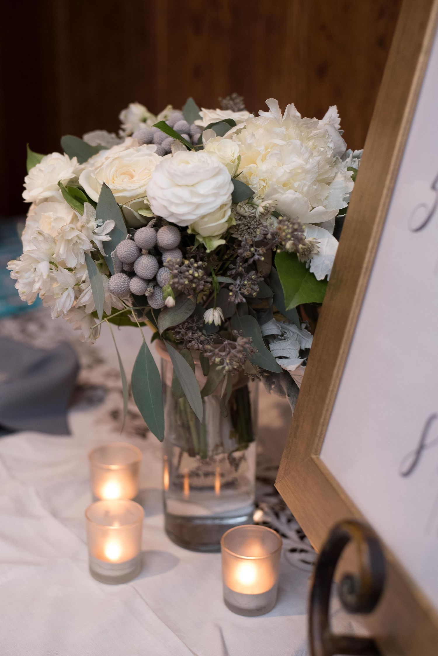 Elegant wedding centerpiece with white roses, carnations, and gray berries in a glass vase, surrounded by glowing votive candles.