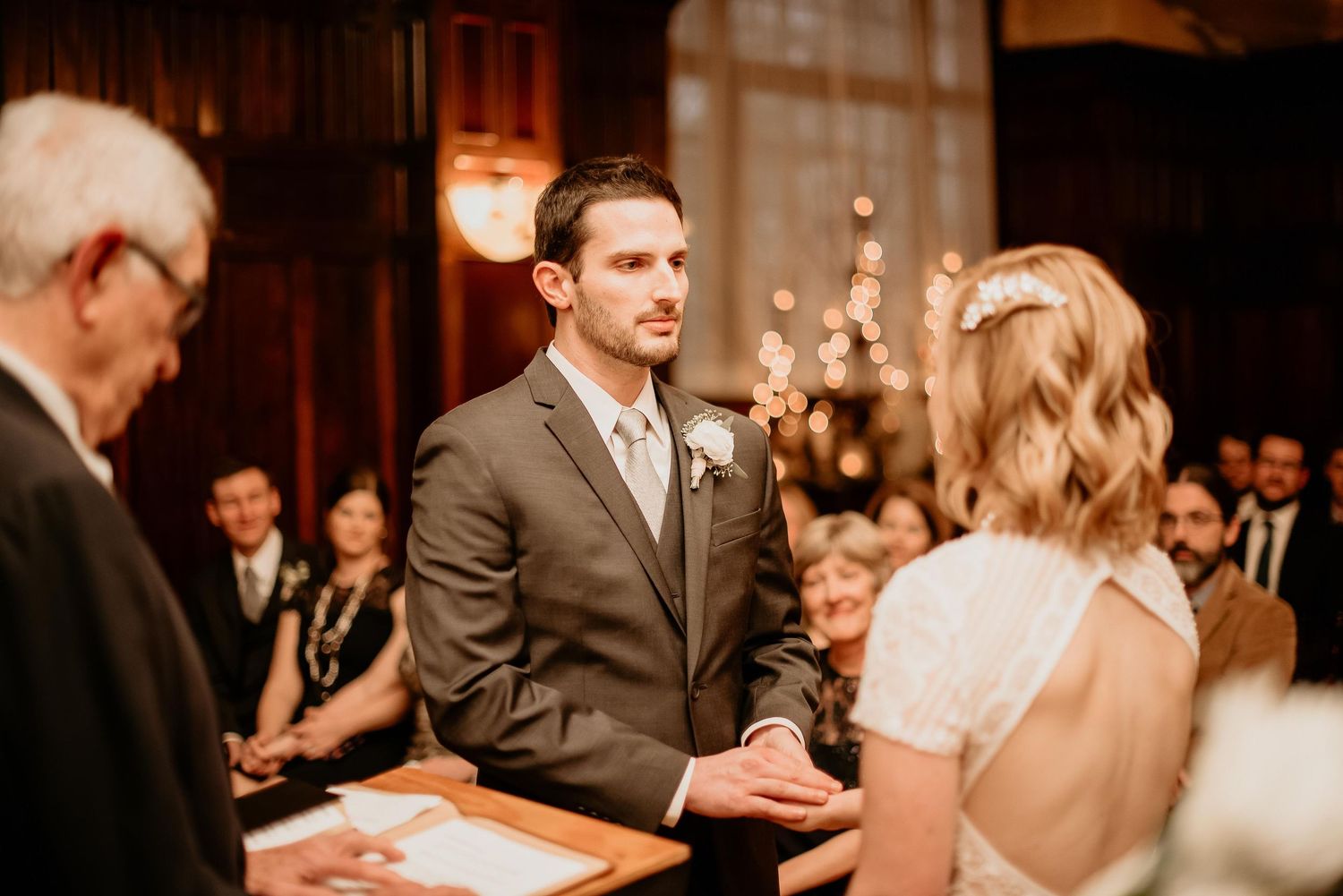 Groom in gray suit exchanges vows with bride during indoor wedding ceremony with warm ambient lighting.
