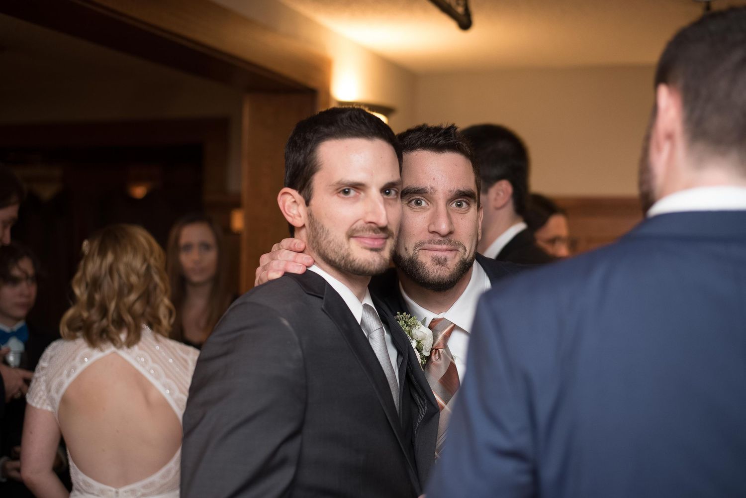 Two men in suits pose together at a wedding reception, smiling at the camera in a warmly lit venue.