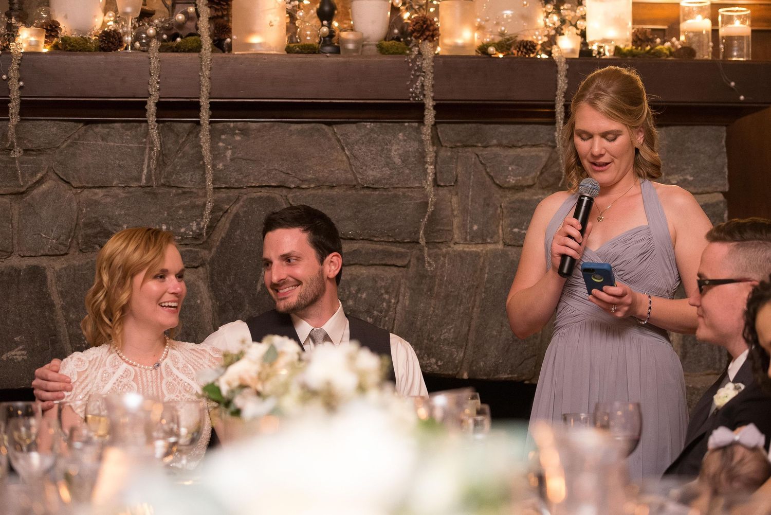 Bridesmaid giving a heartfelt speech at a wedding reception while bride and groom smile at a decorated table.