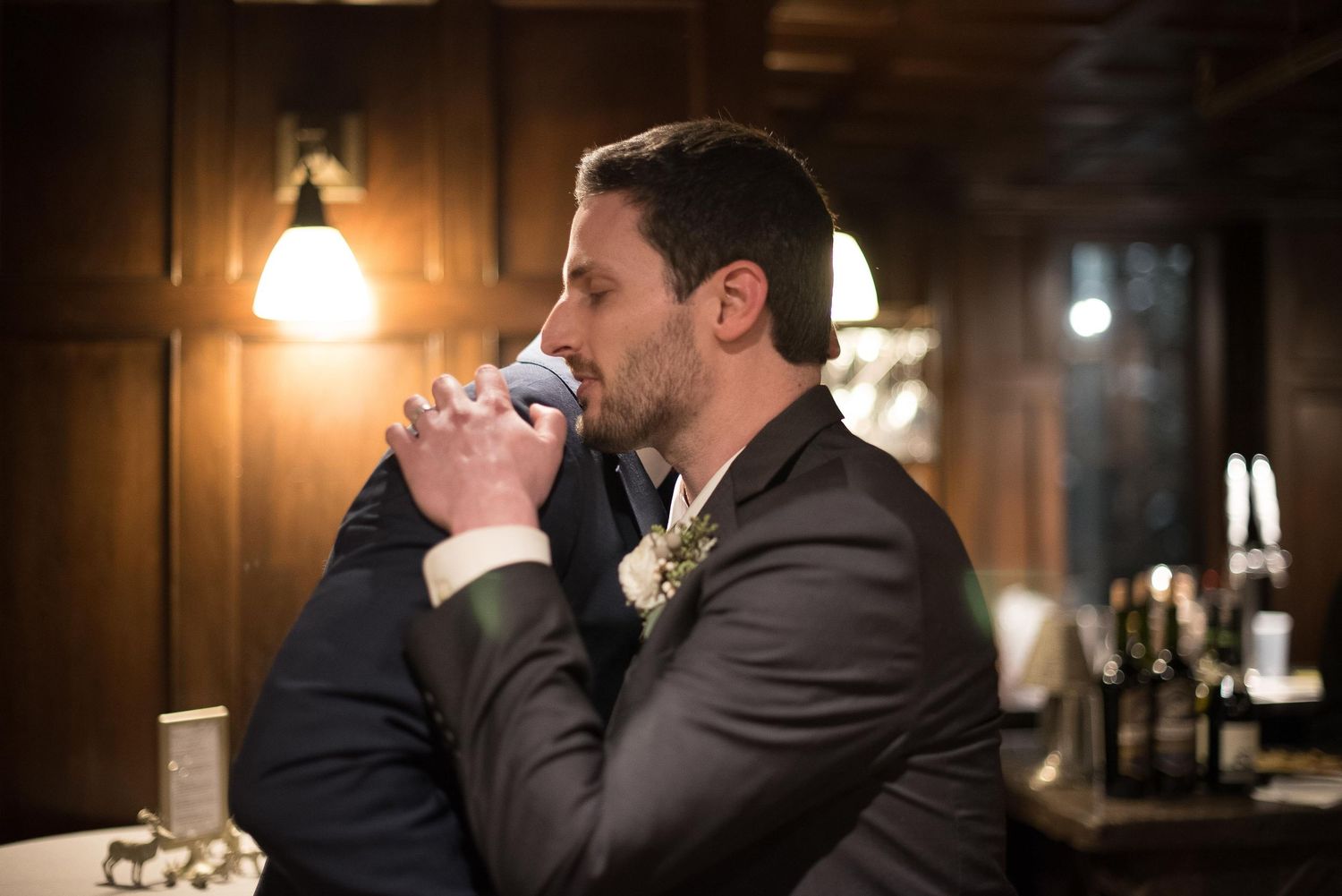 Groom in a brown suit drinking from a glass at a bar during a wedding reception in a wood-paneled venue.