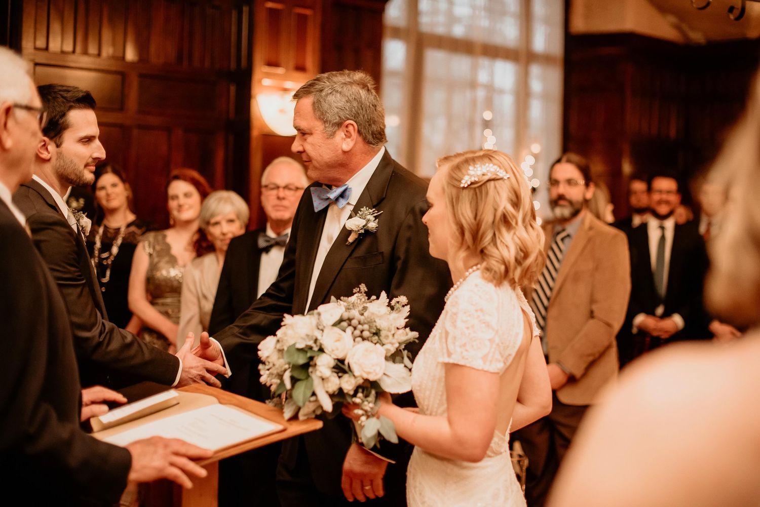 Bride holding white bouquet exchanges vows with groom at elegant indoor wedding ceremony with guests watching.