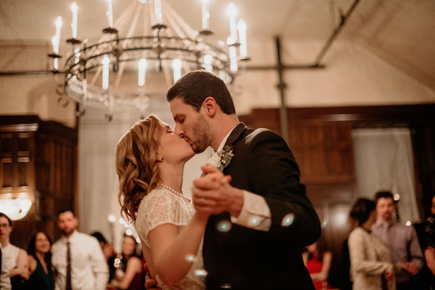 Bride and groom share first dance kiss under elegant chandelier at romantic wedding reception.