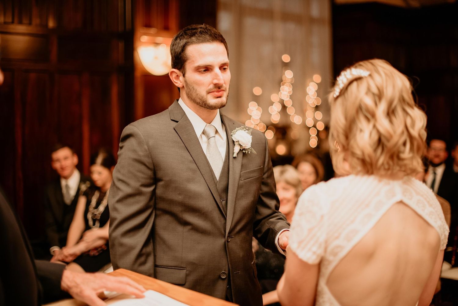 Groom in gray suit holds bride's hands during indoor wedding ceremony with guests seated in background.