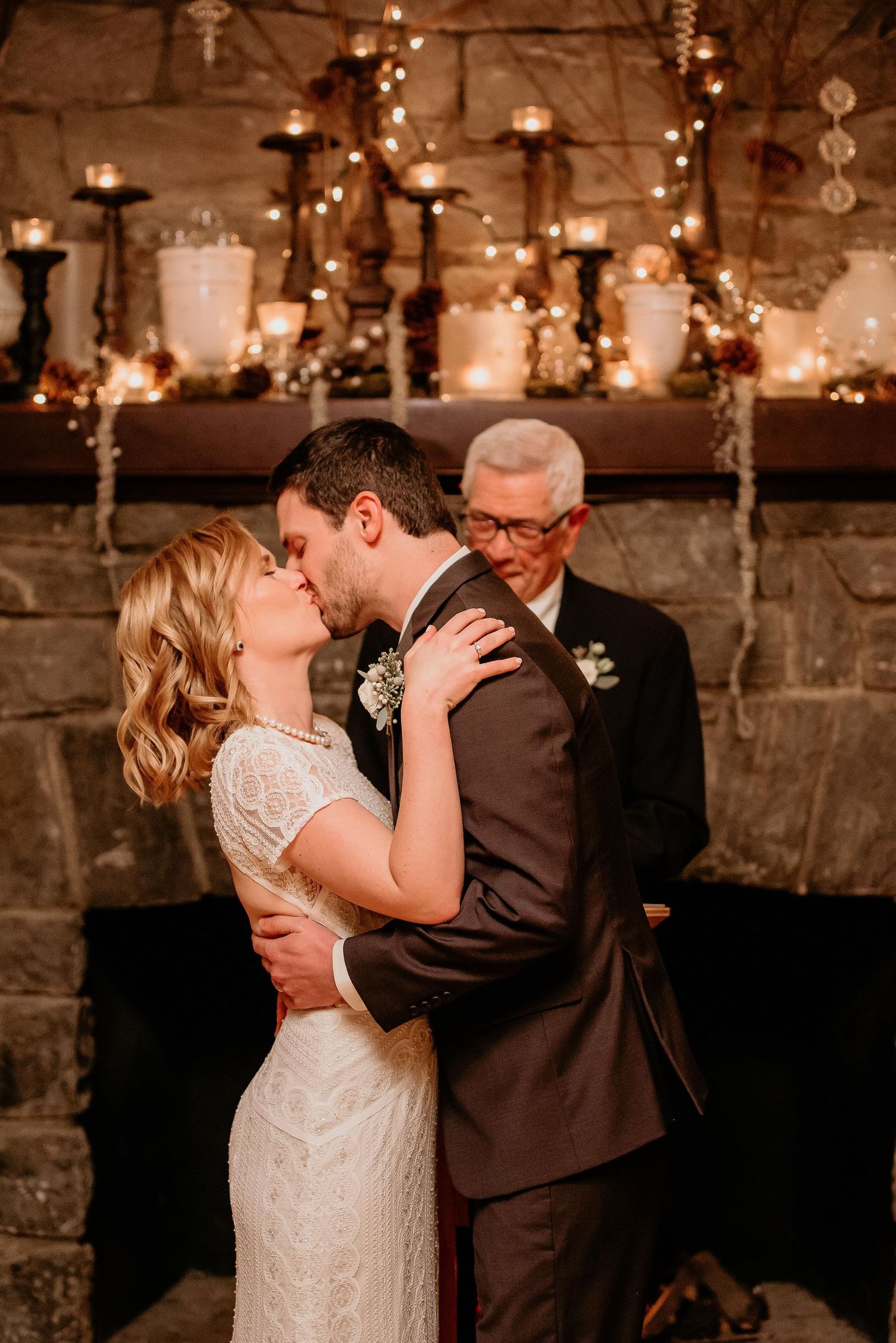 Bride and groom share first kiss at stone fireplace altar adorned with candles and fairy lights at romantic wedding.