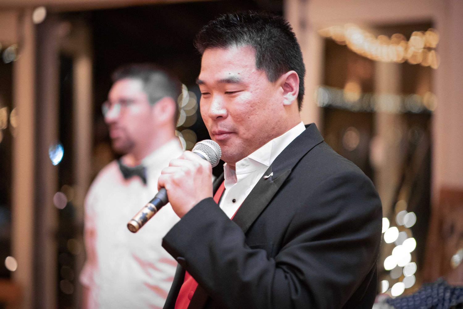 Asian man in black suit singing into microphone at formal event with guests in background.
