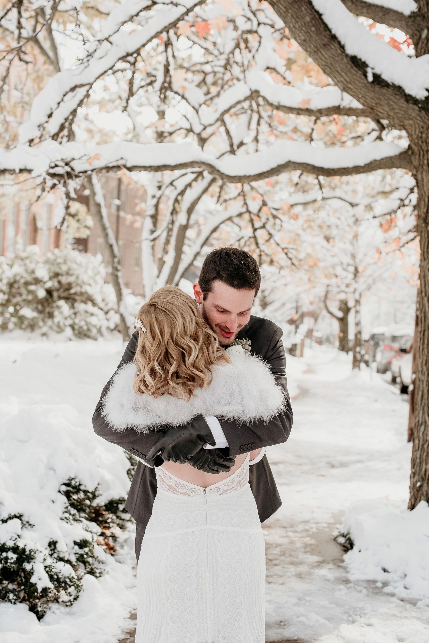 A couple embraces in a snowy winter wonderland, the bride in a white lace gown with fur wrap, groom in a gray suit.