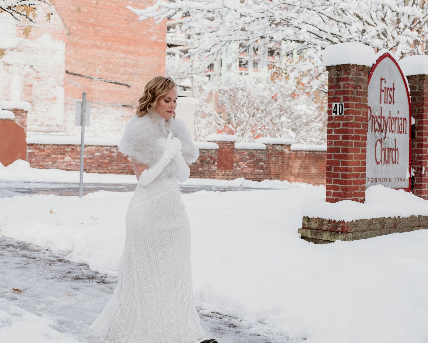 Bride in white lace gown and fur stole standing in snow outside First Presbyterian Church in winter.