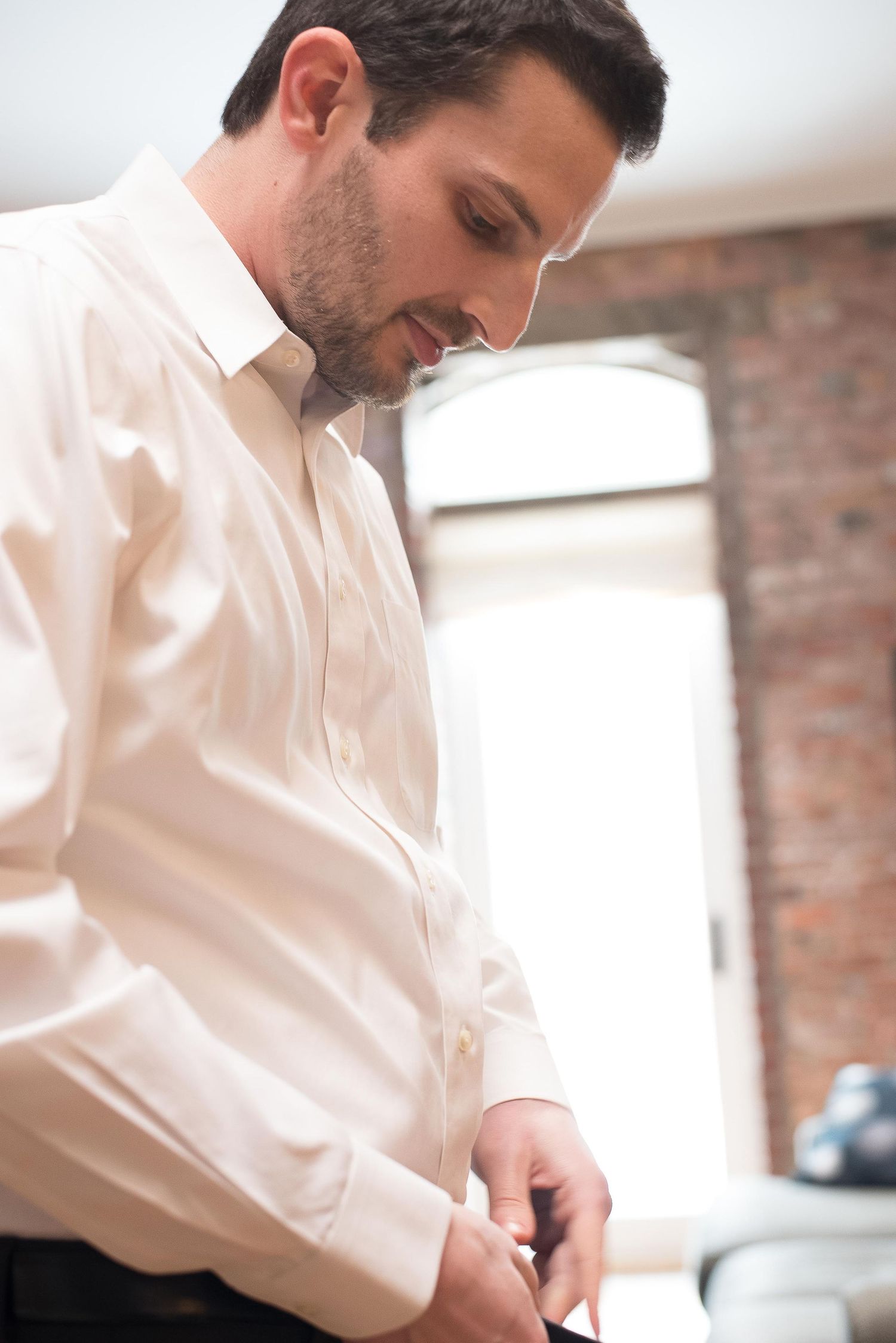 Man in white dress shirt fastening his cuff links while getting ready in a room with brick walls.