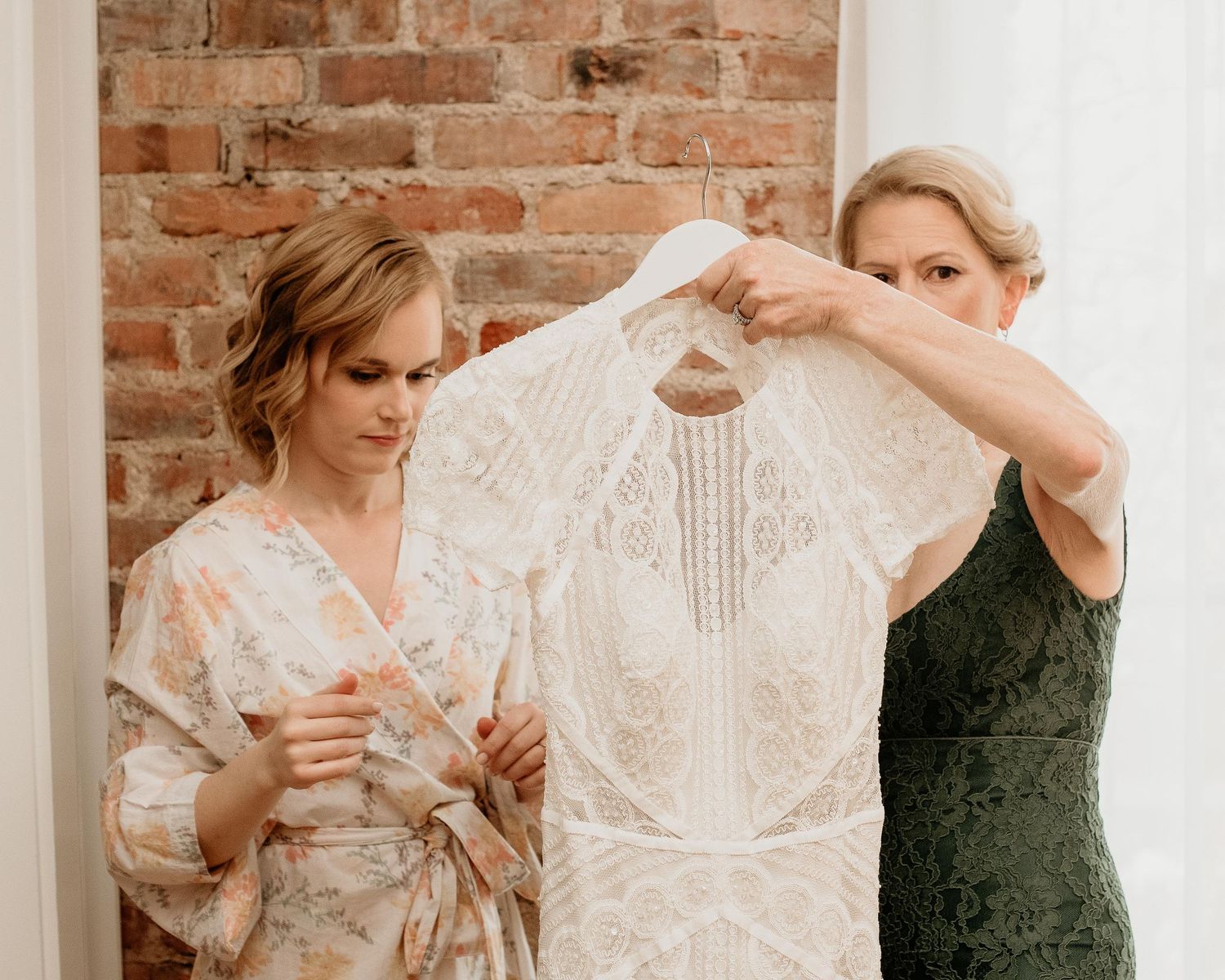 Bride in floral robe and woman in green dress holding up an intricate lace wedding gown near a brick wall.