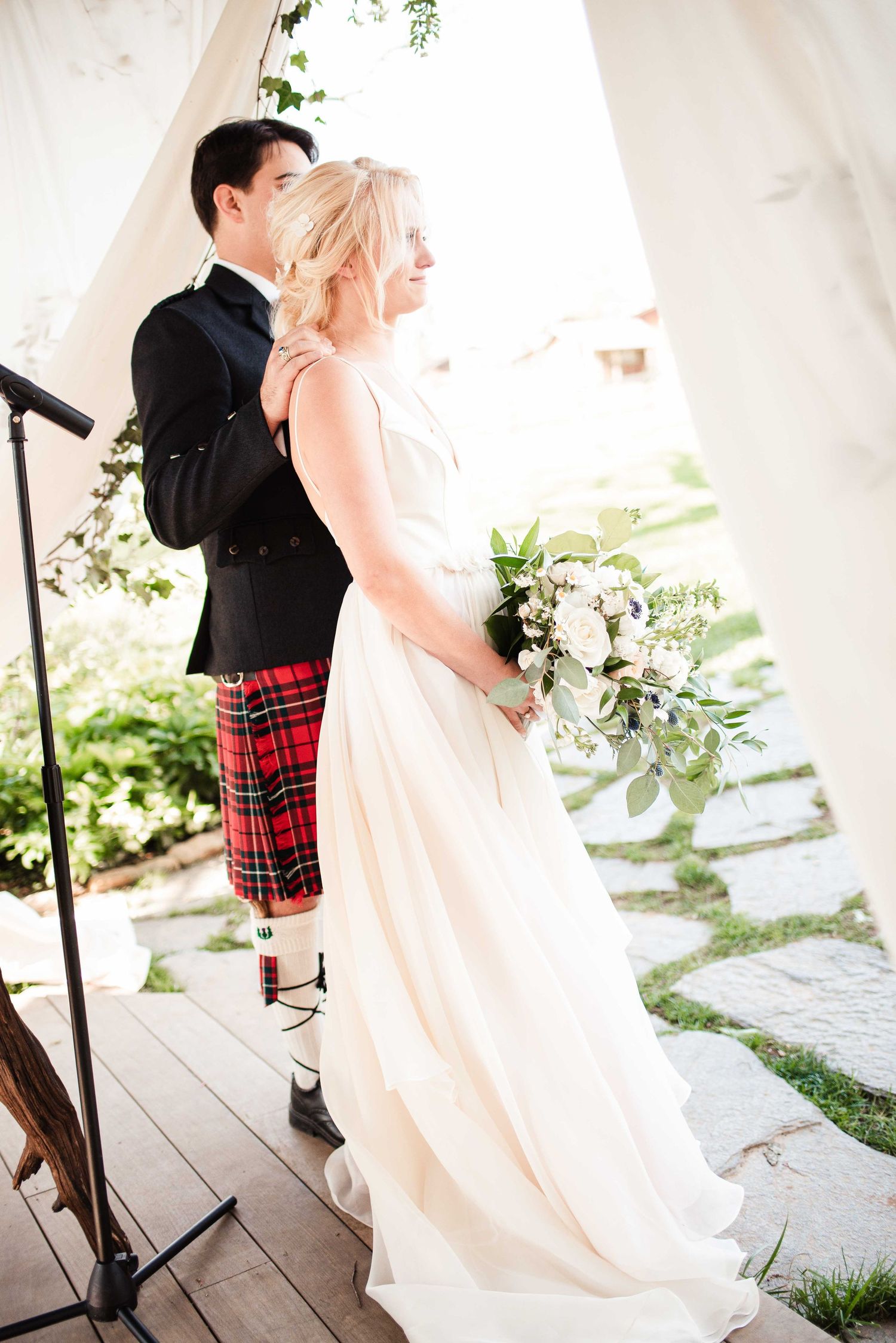 Bride in white gown holding bouquet stands with groom in black jacket and red tartan kilt at outdoor wedding ceremony.