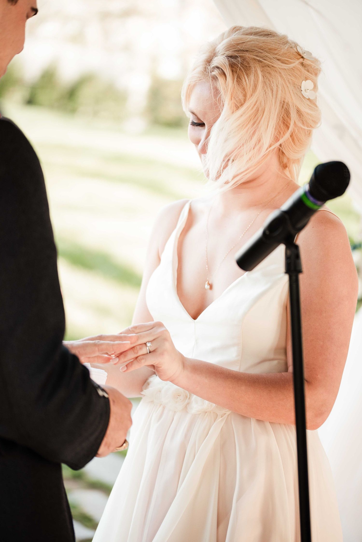 Bride in white gown placing wedding ring on groom's finger during outdoor ceremony near microphone.