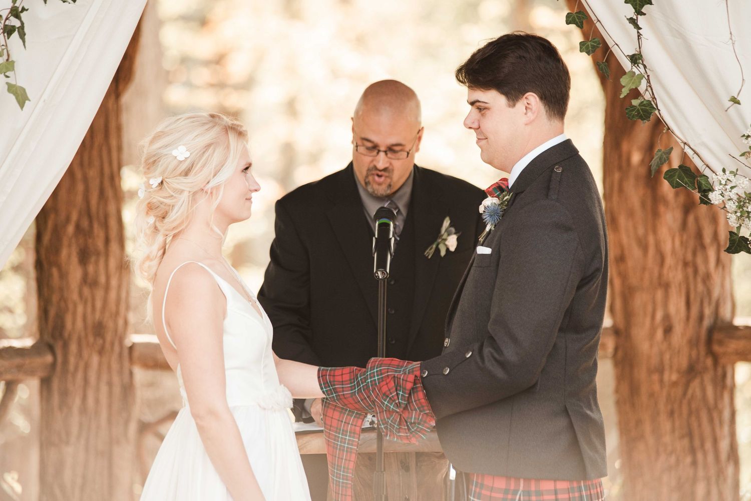 Bride and groom exchange vows outdoors at rustic wedding ceremony with officiant and floral arch backdrop.