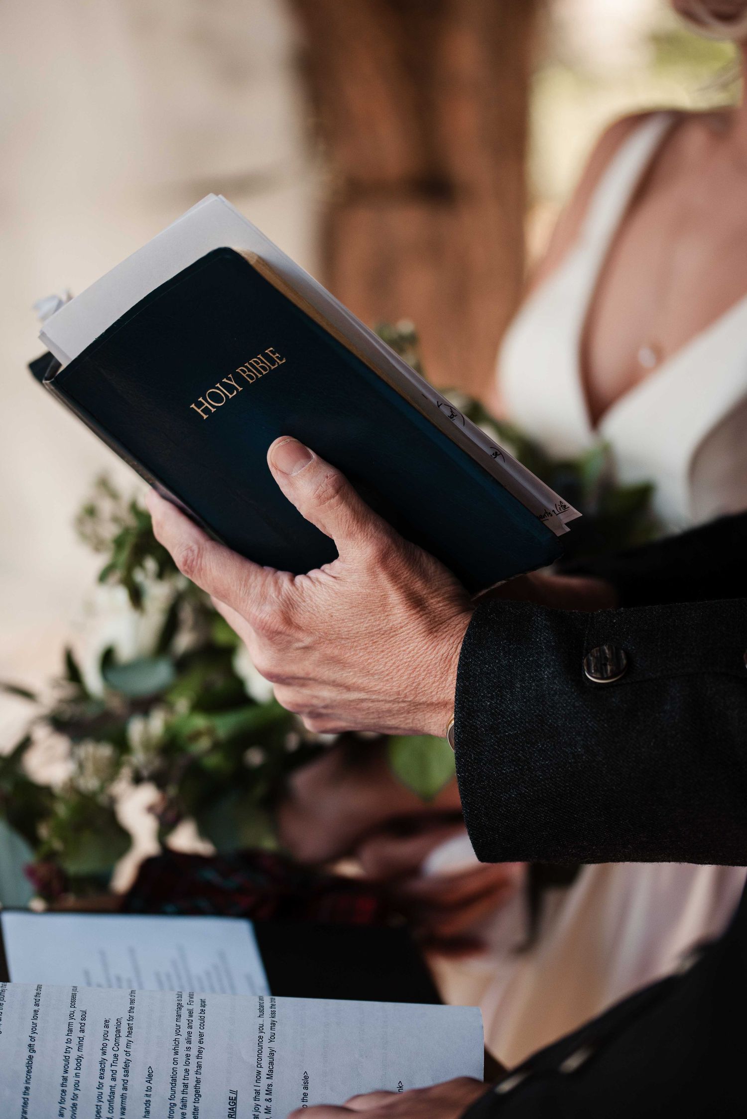 Officiant holding a Holy Bible during an outdoor wedding ceremony with bride holding floral bouquet in background.