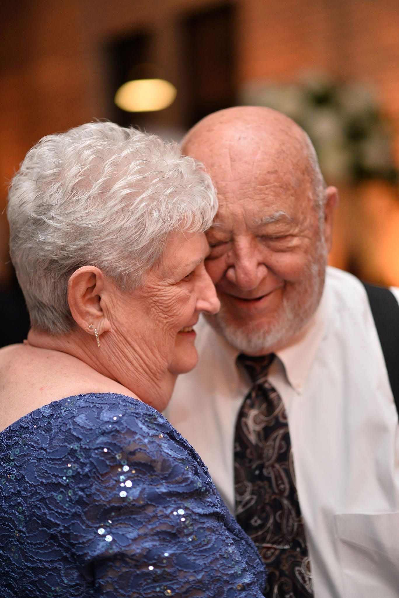 Elderly couple shares a tender forehead-to-forehead moment while dancing at a wedding reception.
