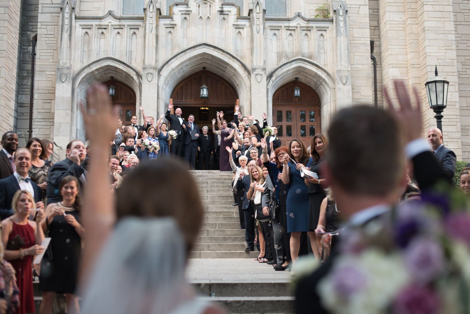 Wedding guests celebrating on steps of grand stone church after ceremony.