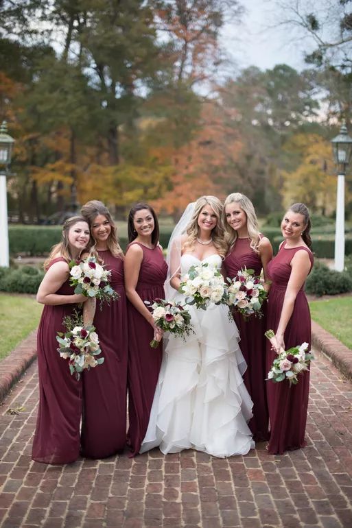 Bride in white gown poses with five bridesmaids in burgundy dresses holding bouquets outdoors.