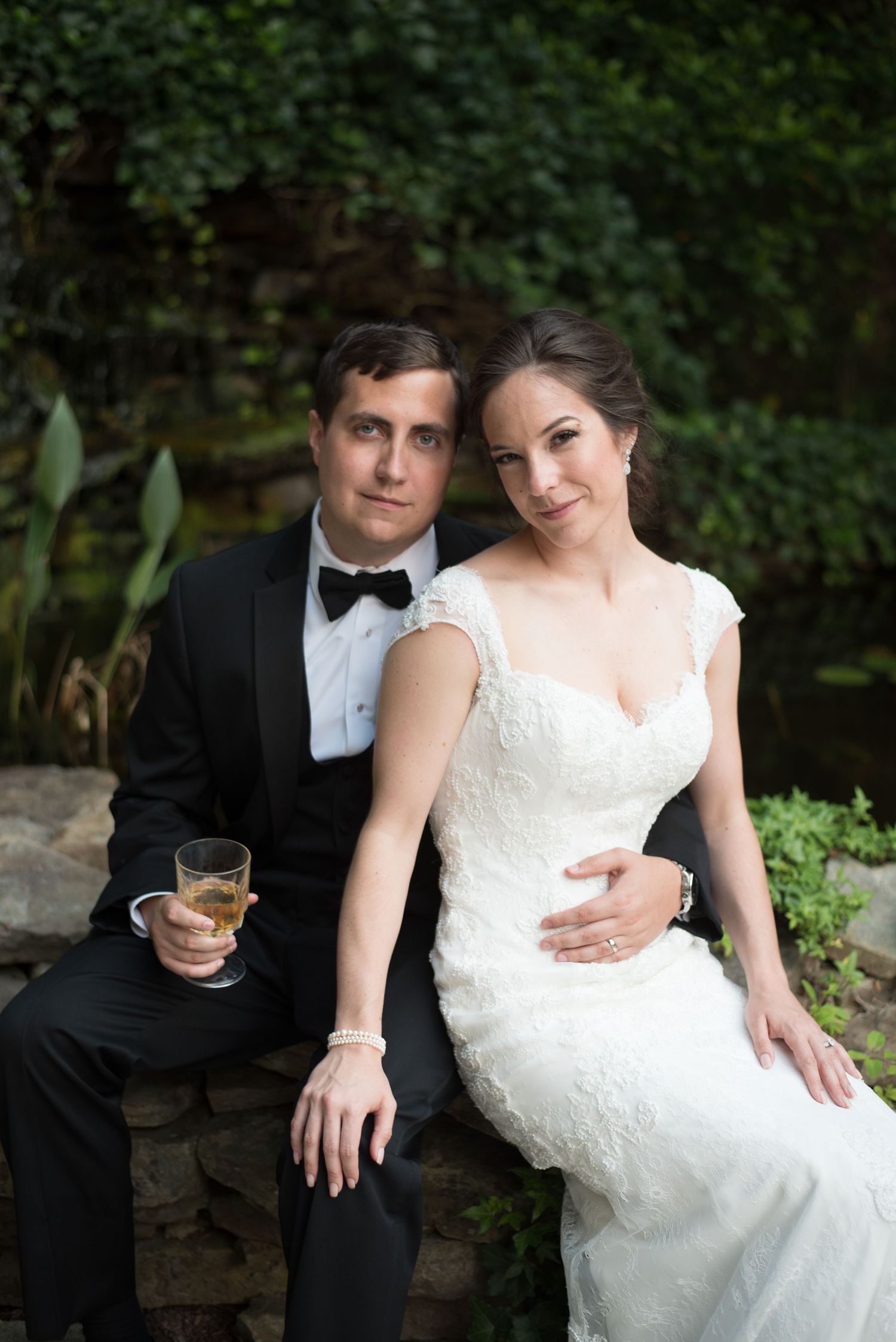 Bride and groom pose together on a stone wall surrounded by lush greenery.