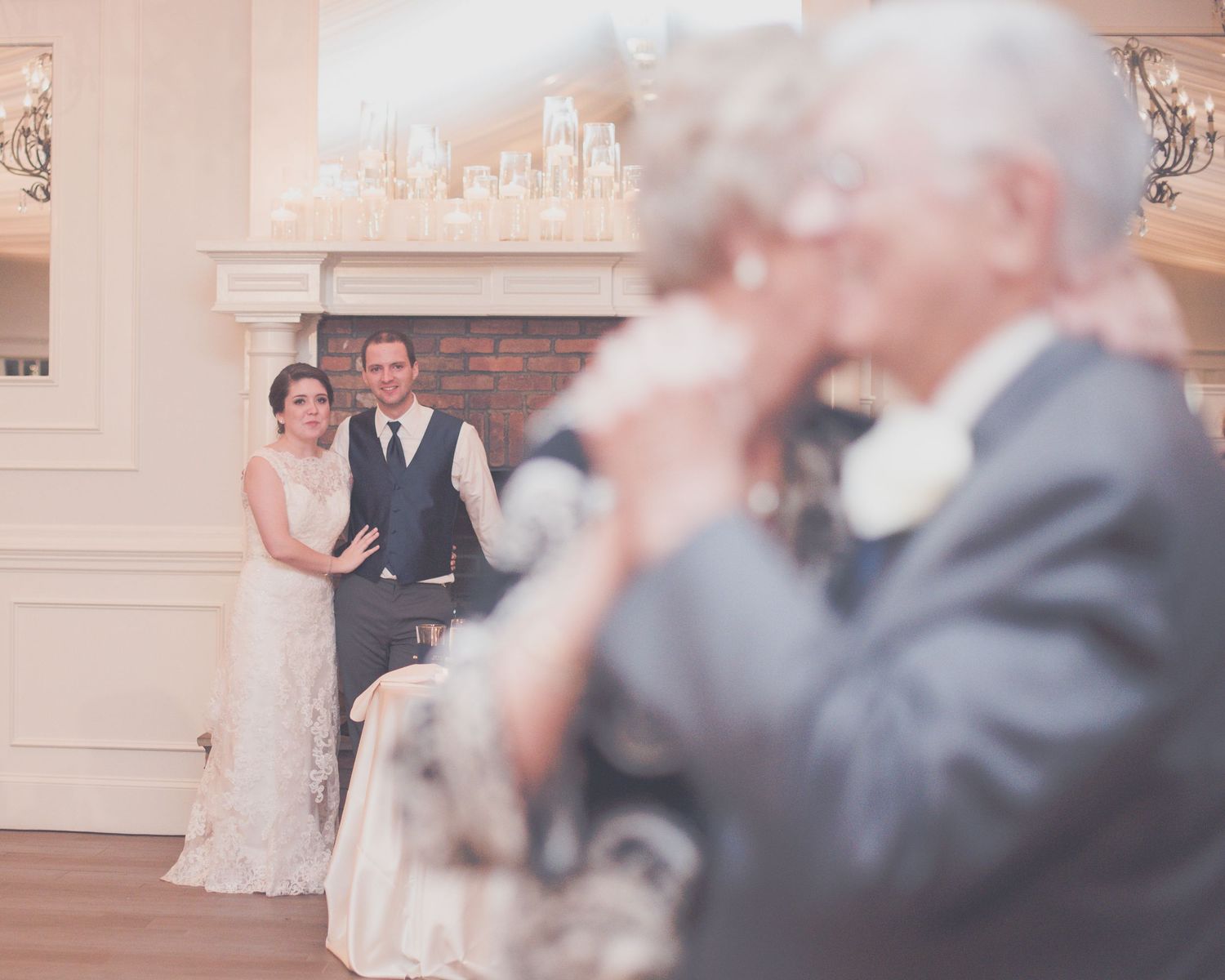 Bride and groom watching elderly couple share a romantic dance near a fireplace at wedding reception.