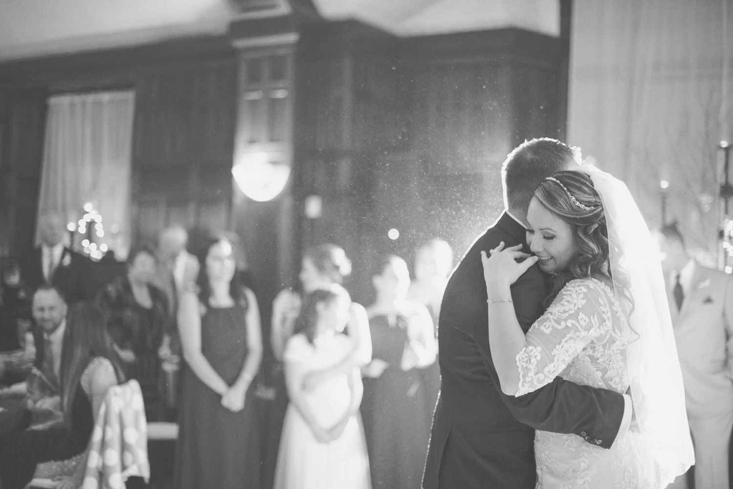 Bride and groom share an emotional first dance in black and white wedding photo with guests watching.