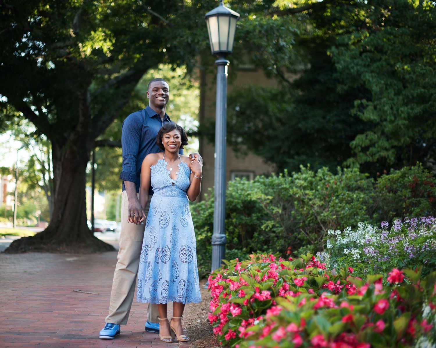 Couple poses together on a flower-lined brick walkway beneath lush green trees during engagement session.
