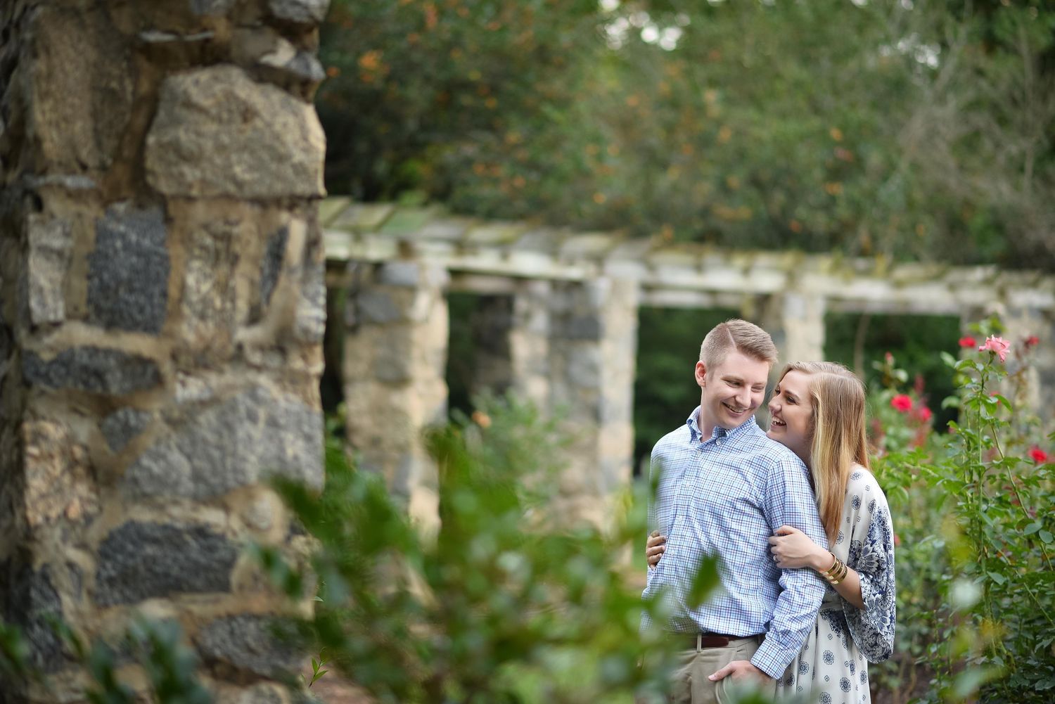 Couple embracing romantically in a lush garden with stone columns and blooming roses in soft natural light.