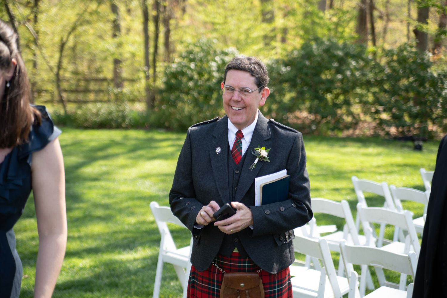 Smiling wedding officiant in traditional Scottish kilt stands in outdoor ceremony venue.