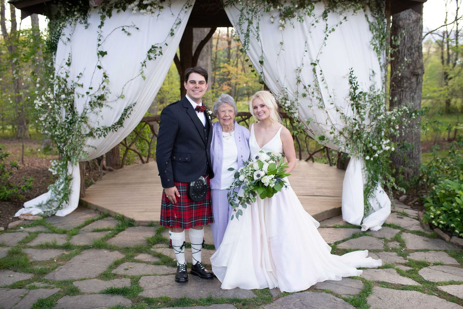 Bride and groom in Scottish kilt posing with elderly woman under floral arch at outdoor wedding ceremony.