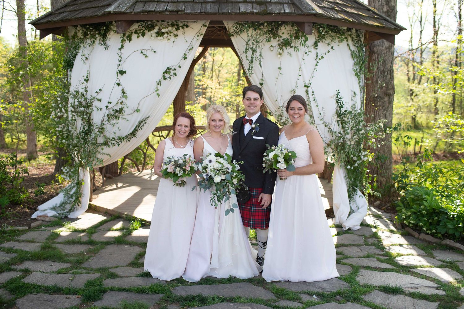 Wedding party poses under floral gazebo, groom in kilt with two bridesmaids in white gowns outdoors.