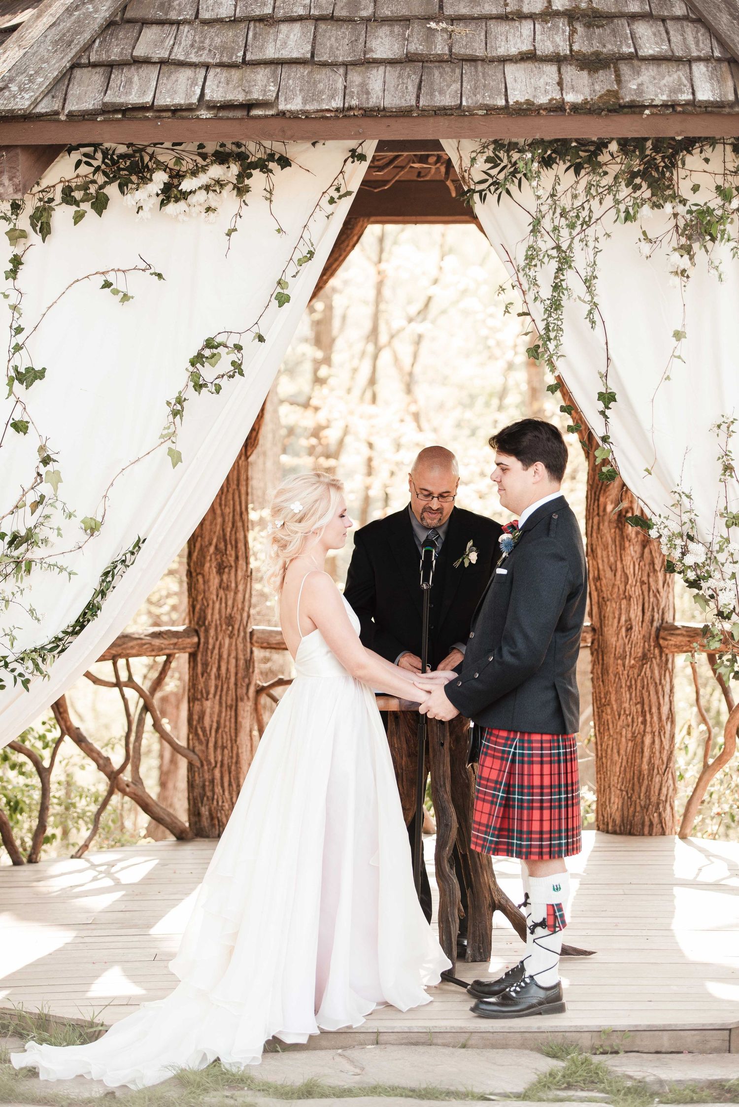 Outdoor wedding ceremony under rustic wooden gazebo with ivy, bride in white gown, groom in Scottish kilt, officiant presiding.