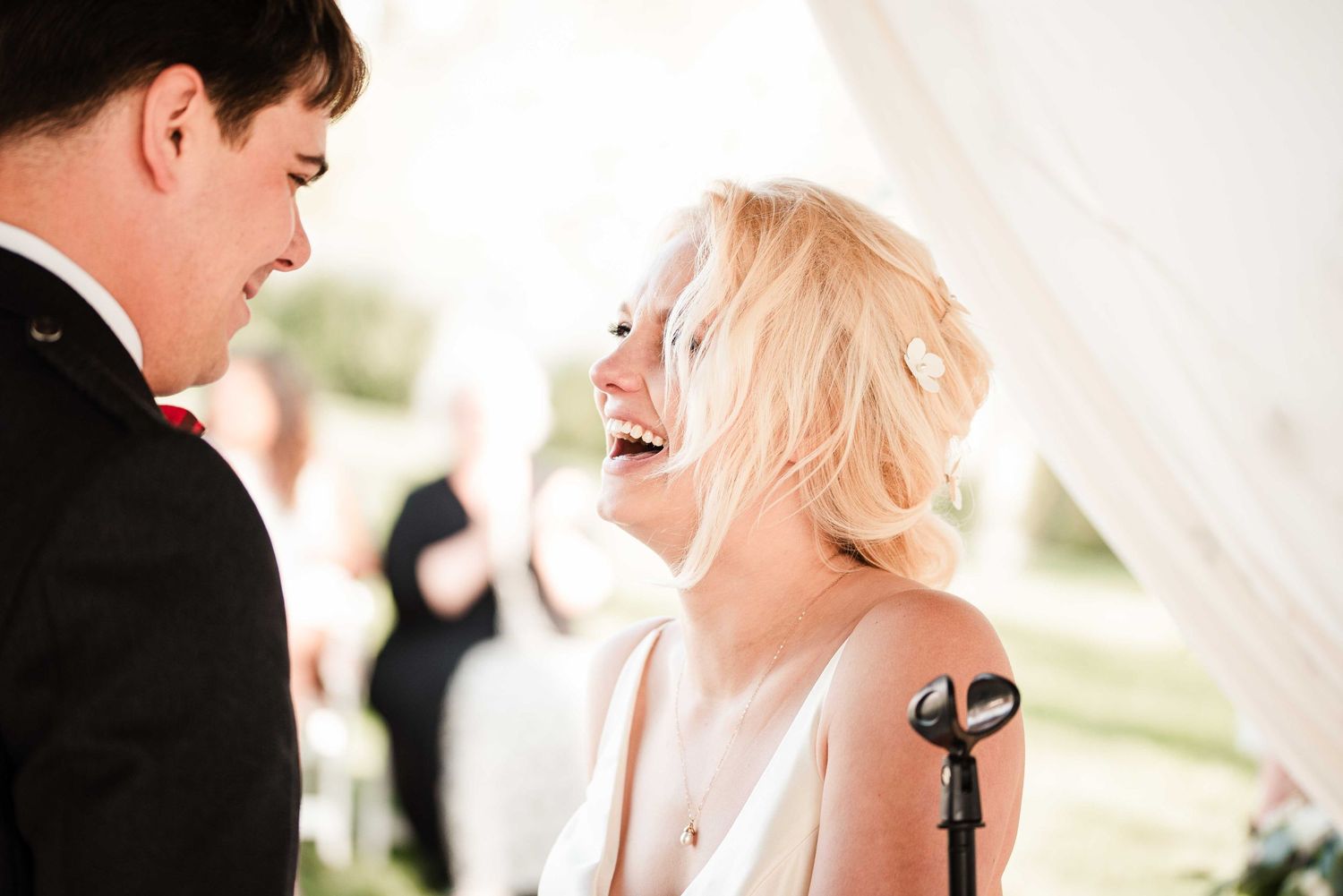 Joyful bride in white dress laughing with groom in black tuxedo at outdoor wedding ceremony.